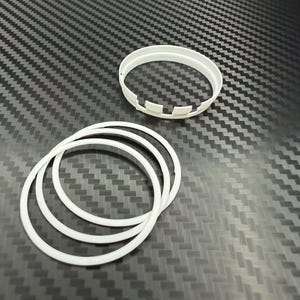 May include: Several white plastic rings and a white plastic ring with notches are arranged on a textured, dark gray surface. The rings appear to be of varying sizes and thicknesses. The background has a repeating pattern.