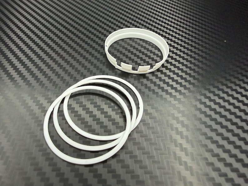 May include: Several white plastic rings and a white plastic ring with notches are arranged on a textured, dark gray surface. The rings appear to be of varying sizes and thicknesses. The background has a repeating pattern.