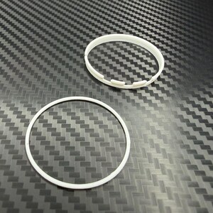 May include: Two white plastic rings on a textured, dark gray surface. One ring is a solid circle, while the other has a series of small, rectangular cutouts along its outer edge. The rings appear to be components of a larger product.