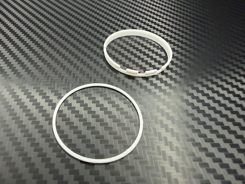 May include: Two white plastic rings on a textured, dark gray surface. One ring is a solid circle, while the other has a series of small, rectangular cutouts along its outer edge. The rings appear to be components of a larger product.