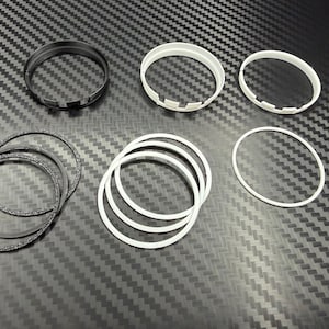 May include: An assortment of black and white circular components, including rings and notched pieces, are arranged on a textured, dark gray surface. The components vary in size and texture, with some appearing smooth and others having a speckled finish.