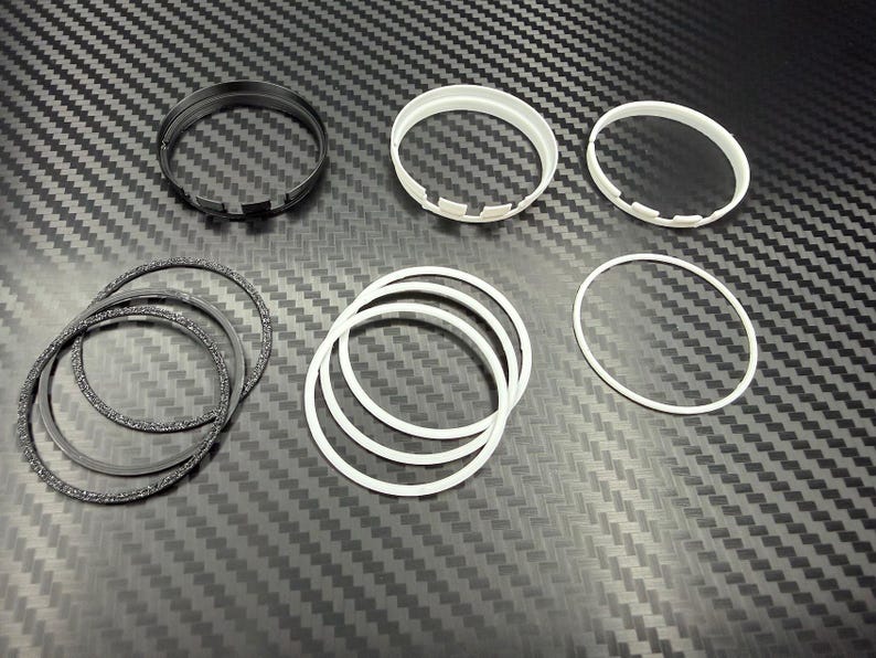 May include: An assortment of black and white circular components, including rings and notched pieces, are arranged on a textured, dark gray surface. The components vary in size and texture, with some appearing smooth and others having a speckled finish.