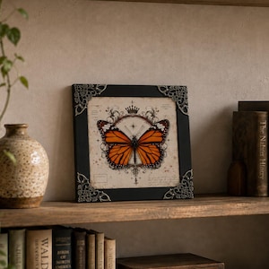 May include: A square black frame with ornate silver-toned metal corner accents containing a print of a monarch butterfly. The butterfly is centered over a vintage-style background featuring a crown icon, starburst, and faint cursive script text.