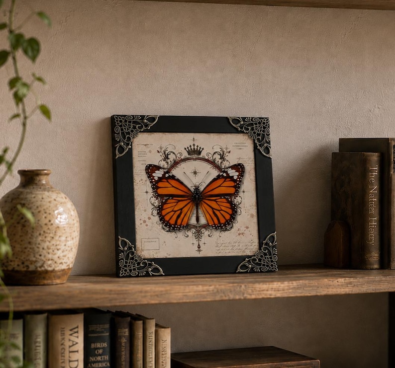 May include: A square black frame with ornate silver-toned metal corner accents containing a print of a monarch butterfly. The butterfly is centered over a vintage-style background featuring a crown icon, starburst, and faint cursive script text.