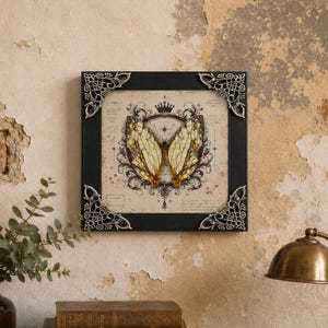 May include: A square wall art piece featuring a cream-colored print of a butterfly centered within a dark, ornate frame. The frame has silver-toned, intricate metal corner accents. The butterfly illustration is surrounded by a crown and scrollwork.