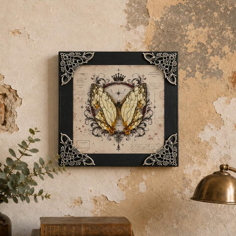 May include: A square wall art piece featuring a cream-colored print of a butterfly centered within a dark, ornate frame. The frame has silver-toned, intricate metal corner accents. The butterfly illustration is surrounded by a crown and scrollwork.