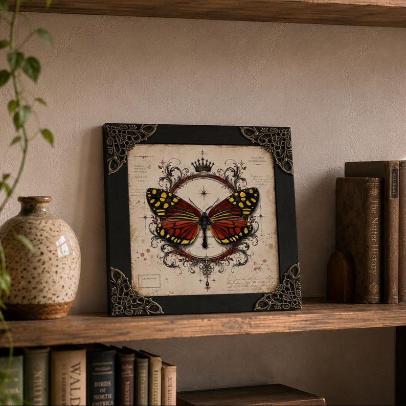 May include: A square framed print featuring a central butterfly with orange and black wings, enclosed in a decorative circular emblem with a crown icon. The background has vintage-style script and markings. The black frame has ornate metal corner accents.