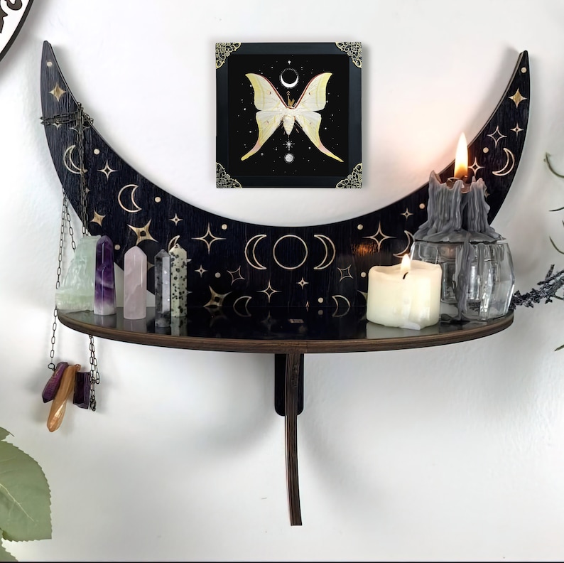 May include: A black crescent moon-shaped wall shelf featuring gold celestial star and moon engravings. The shelf holds several crystal points, a chain with hanging stones, and two candles, with a framed moth print above it.