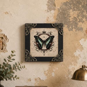 May include: A square wall art piece featuring a black frame with ornate metallic corner accents. The central print displays a teal and black butterfly centered within a circular decorative emblem topped with a crown, set against a beige background.