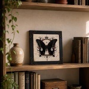 May include: A square black frame with ornate corner accents containing a print of a large black butterfly centered over a vintage-style map background with a crown icon, decorative scrollwork, and fine lines radiating from the center point.
