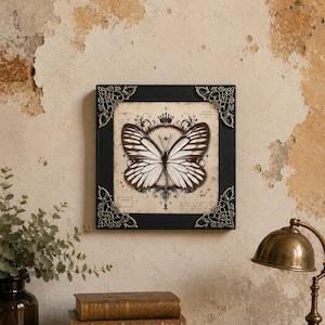 May include: A square wall art piece featuring a central illustration of a white and brown butterfly with striped wings. The artwork is set within a black frame adorned with ornate, metallic silver-toned corner accents and a crown motif.