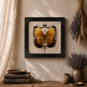 May include: A square framed wall art piece featuring a large, golden-yellow butterfly with dark brown wing edges. The butterfly is centered over a vintage-style background with a crown icon, compass-like markings, and faint cursive script text.