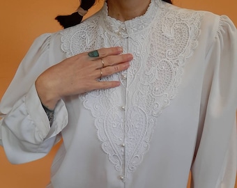 Könnte beinhalten: Eine weiße Bluse mit hohem, gerüschtem Kragen und langen Ärmeln. Die Vorderseite hat eine detaillierte, weiße Spitzentafel und perlenartige Knöpfe. Die Bluse hat eine Vintage-Ästhetik.