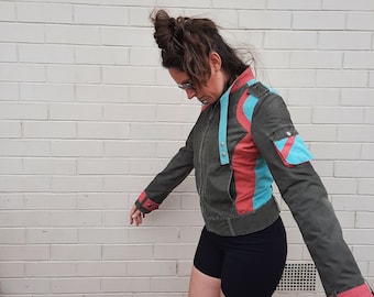 Könnte beinhalten: Eine grüne, rosa und blaue Jacke mit Kapuze. Die Jacke hat einen Reißverschluss und eine aufgesetzte Tasche auf der linken Seite. Das Model trägt eine schwarze Shorts.