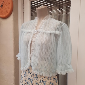 May include: A light blue sheer cardigan with a ruffled trim and a tie at the neck. The cardigan is on a mannequin and is displayed against a white background.