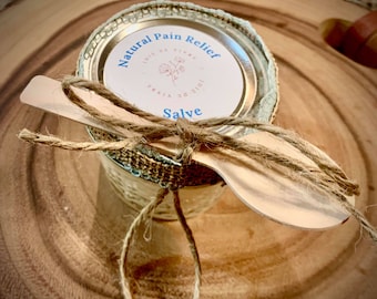 以下が含まれることがあります： 「Natural Pain Relief」と「Joie de Vivre」の文字が入った蓋が付いた、天然の鎮痛軟膏の瓶。瓶は麻ひもで包まれ、木のスプーンが上に置かれています。