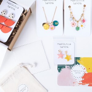 May include: A flat lay image of a small business's packaging for jewelry. The image shows a variety of jewelry pieces, including necklaces, earrings, and charms. The jewelry is packaged in small white boxes with the business's name, "MARIQUILLA SANDIA", printed on them. The jewelry is also packaged in a small white drawstring bag with the business's name printed on it.