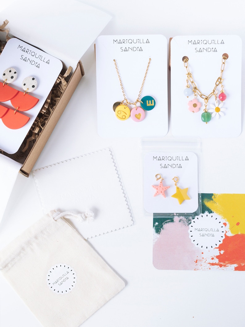 May include: A flat lay image of a small business's packaging for jewelry. The image shows a variety of jewelry pieces, including necklaces, earrings, and charms. The jewelry is packaged in small white boxes with the business's name, "MARIQUILLA SANDIA", printed on them. The jewelry is also packaged in a small white drawstring bag with the business's name printed on it.