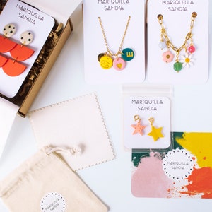May include: A set of colorful jewelry pieces, including a necklace with a heart, a star, and a letter charm, a necklace with a flower and a daisy charm, and a pair of earrings with a watermelon and polka dot design. The jewelry is packaged in a small box with a white bag and a white card with the text "MARIQUILLA SANDIA".