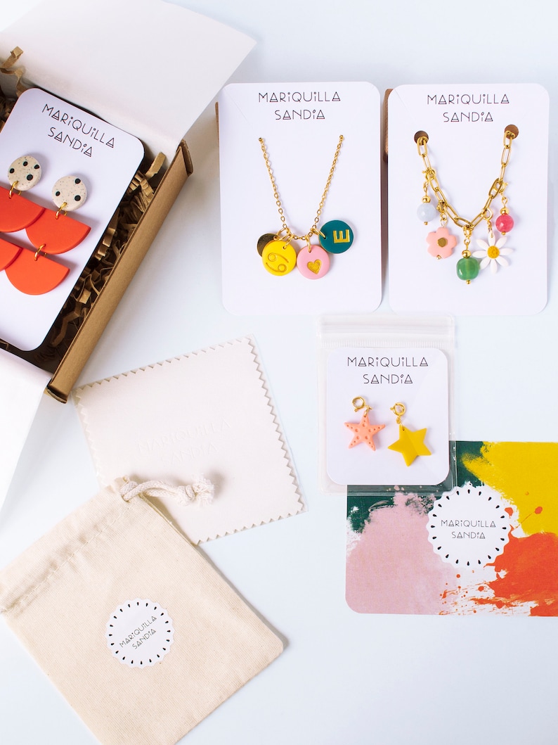 May include: A set of colorful jewelry pieces, including a necklace with a heart, a star, and a letter charm, a necklace with a flower and a daisy charm, and a pair of earrings with a watermelon and polka dot design. The jewelry is packaged in a small box with a white bag and a white card with the text "MARIQUILLA SANDIA".