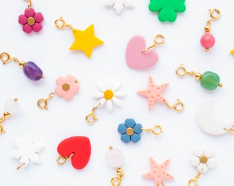 Puede incluir: Una colección de abalorios coloridos, que incluyen un corazón rosa, una estrella amarilla, un trébol verde, una flor rosa, una flor azul, una flor blanca, una estrella de mar rosa, un copo de nieve blanco, una luna creciente blanca y una estrella de mar rosa y blanca a rayas. Los abalorios están hechos de arcilla polimérica y tienen anillas de salto doradas.