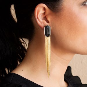 Può includere: Orecchini dorati con una pietra nera ovale e frange a cascata. Questi orecchini lunghi e pendenti offrono un look glamour. Il design è semplice ma elegante, adatto a varie occasioni. Un accessorio di moda.