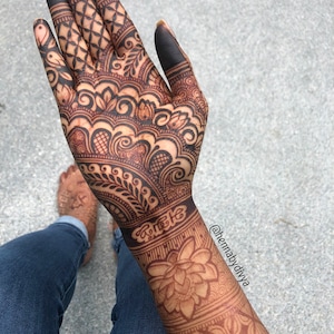 May include: Intricate henna design on a hand and forearm, featuring a large floral motif and geometric patterns. The design is dark brown and covers the entire hand and forearm. The person is wearing blue jeans.
