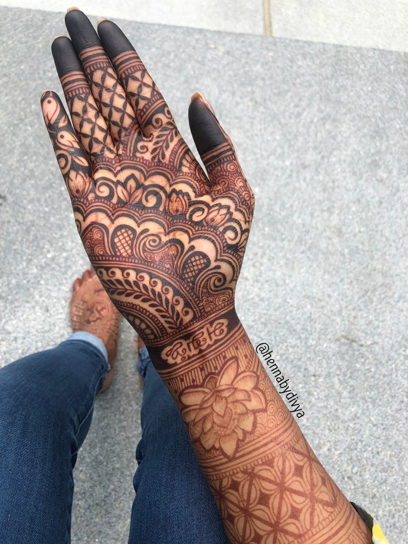 May include: Intricate henna design on a hand and forearm, featuring a large floral motif and geometric patterns. The design is dark brown and covers the entire hand and forearm. The person is wearing blue jeans.