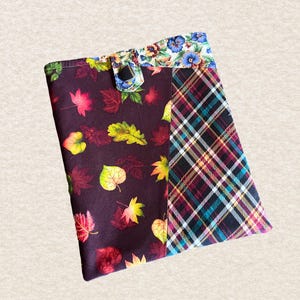 May include: A rectangular fabric pouch featuring a dark brown section with a colorful autumn leaf print and a black, white, and teal plaid section. The top flap is secured with a small tab and features a floral pattern in blue and green tones.