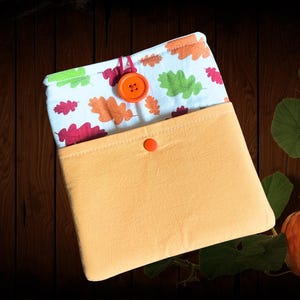 Può includere: Una custodia in tessuto con esterno in tinta unita arancione chiaro e una chiusura a pressione arancione. La fodera interna è in tessuto bianco con un motivo di piccole foglie di quercia verdi, arancioni e rosso scuro, con un bottone.