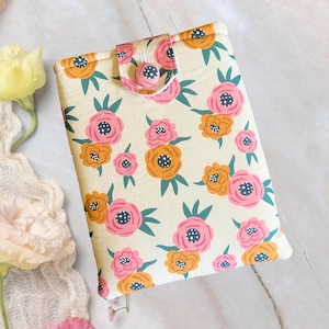 May include: A floral book cover with a cream background and a pattern of pink and orange flowers with green leaves. The cover has a button closure and a matching floral design. The cover is approximately 9 inches long and 6 inches wide.