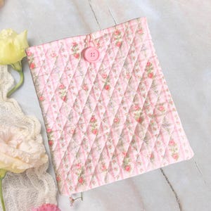 Op de afbeelding: Een roze gewatteerde boekomslag met een bloemenpatroon en een knoopsluiting. De omslag heeft een ruitpatroon en is versierd met kleine roze en rode rozen. De achtergrond bevat kant en bloemen.