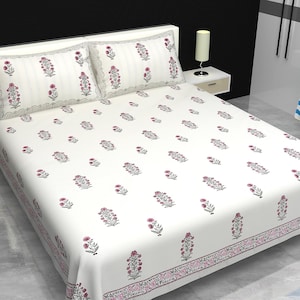 Puede incluir: Una colcha blanca y dos fundas de almohada con un estampado floral en rosa y gris. La colcha tiene un borde con un diseño similar. Las almohadas tienen un borde festoneado.