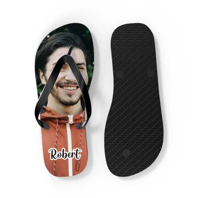May include: A pair of flip-flops featuring a black rubber strap. The left sandal displays a photographic print of a smiling person wearing an orange jacket, with the name Robert printed in white script. The right sandal has a textured black sole.