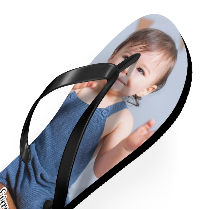 May include: A flip-flop featuring a custom photographic print of a young child wearing a blue romper. The footwear has a solid black rubber sole and a matching black Y-shaped strap that secures the foot. The image of the child covers the footbed.