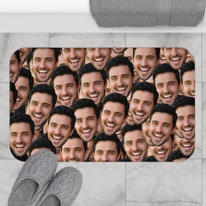 May include: A rectangular bath mat featuring a repeating pattern of a smiling man's face printed across the entire surface. The mat has rounded corners and is positioned on a light grey tiled floor with a grey textured towel visible at the top.