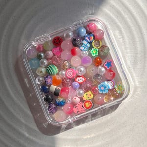 May include: A clear plastic container filled with colourful beads and charms. The beads are a variety of shapes and sizes, including round, star-shaped, and heart-shaped. The charms are also a variety of shapes and sizes, including smiley faces, flowers, and animals. The beads and charms are a mix of pink, purple, blue, green, yellow, and red.