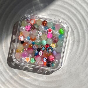 May include: A clear plastic container filled with colourful beads, charms, and glitter. The beads are in various shapes, sizes, and colours, including pink, blue, green, yellow, and white. Some of the charms include stars, hearts, and a seashell.