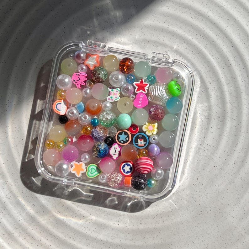 May include: A clear plastic container filled with colourful beads, charms, and glitter. The beads are in various shapes, sizes, and colours, including pink, blue, green, yellow, and white. Some of the charms include stars, hearts, and a seashell.