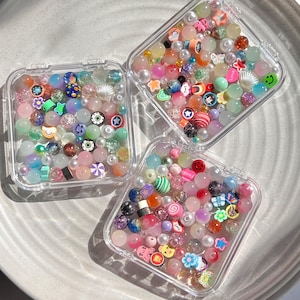 May include: Three clear plastic containers filled with colourful beads and charms. The beads are a mix of pastel colours, clear, and opaque. The charms are small and include smiley faces, stars, and other shapes.