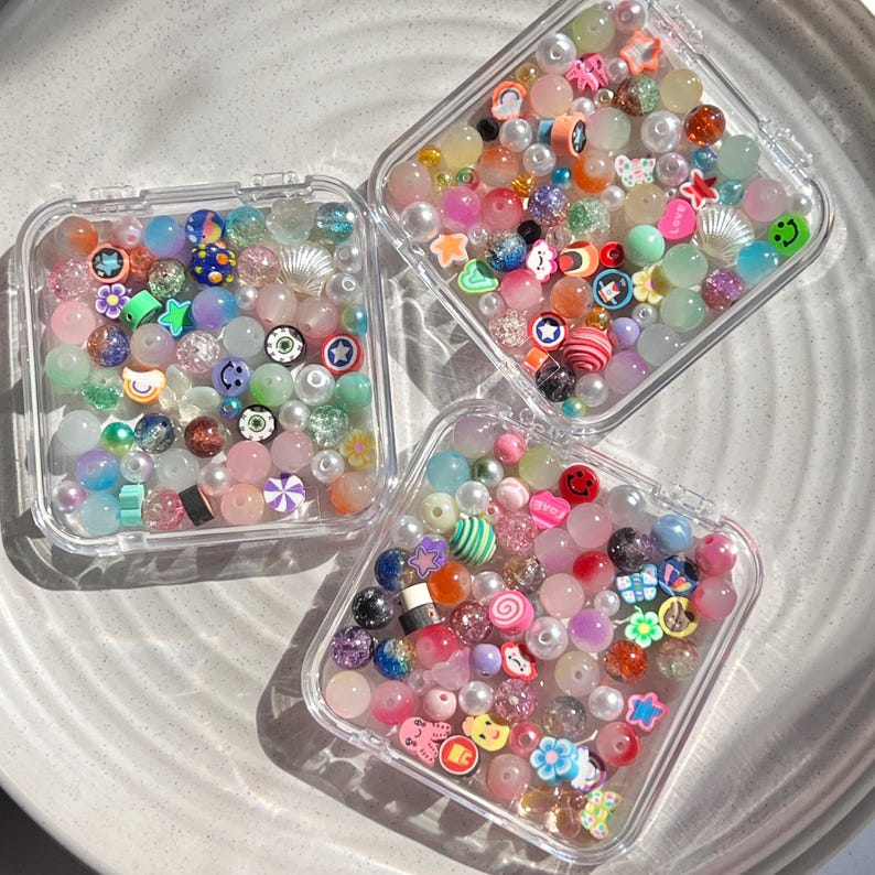 May include: Three clear plastic containers filled with colourful beads and charms. The beads are a mix of pastel colours, clear, and opaque. The charms are small and include smiley faces, stars, and other shapes.