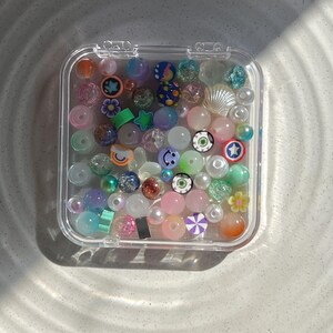 May include: A clear plastic container filled with colourful beads. The beads are a variety of shapes and sizes, including round, square, and star-shaped. Some of the beads have glitter or other decorative elements.