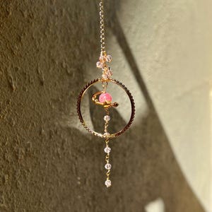 May include: A gold chain necklace with a pink gemstone pendant inside a circular frame. The pendant is suspended from a smaller gold chain. The necklace is adorned with small clear crystals.