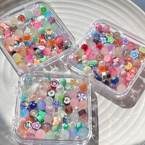May include: Three clear plastic containers filled with colorful beads and charms. The beads are a mix of pastel colors, including pink, blue, green, and yellow. The charms are small and include shapes like stars, hearts, and smiley faces.