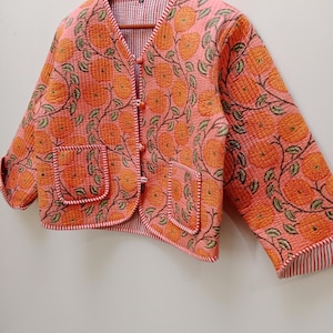 Op de afbeelding: Een roze gewatteerd jack met een bloemenprint en knoopsluiting. Het jack heeft twee zakken en een gestreepte voering.