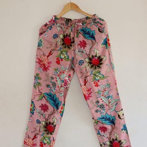 May include: Pink floral pajama trousers with a drawstring waist, hanging on a wooden hanger. The pattern features red, blue, green, and brown flowers and leaves. The trousers are designed for comfort and relaxation.