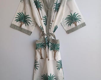 May include: White robe with a green and brown palm tree pattern. The robe has a striped trim and a matching belt. The sleeves and edges are also trimmed with green stripes. The robe is hanging on a wooden hanger.