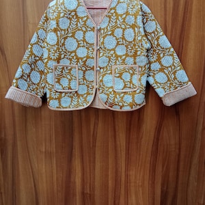 May include: A mustard yellow quilted jacket with a white floral print and a button closure. The jacket has two pockets and a striped trim on the sleeves.