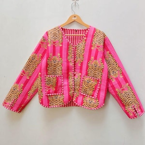 Puede incluir: Chaqueta acolchada rosa con cuello en V y mangas largas. La chaqueta presenta un patrón de rayas verticales con detalles de estampado de tigre y dos bolsillos delanteros. La chaqueta está colgada en una percha de madera.