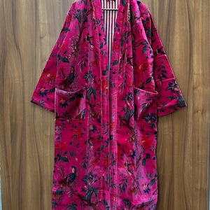 May include: A long pink velvet robe with a floral print featuring birds and leaves. The robe has a white and red striped lining and two pockets.